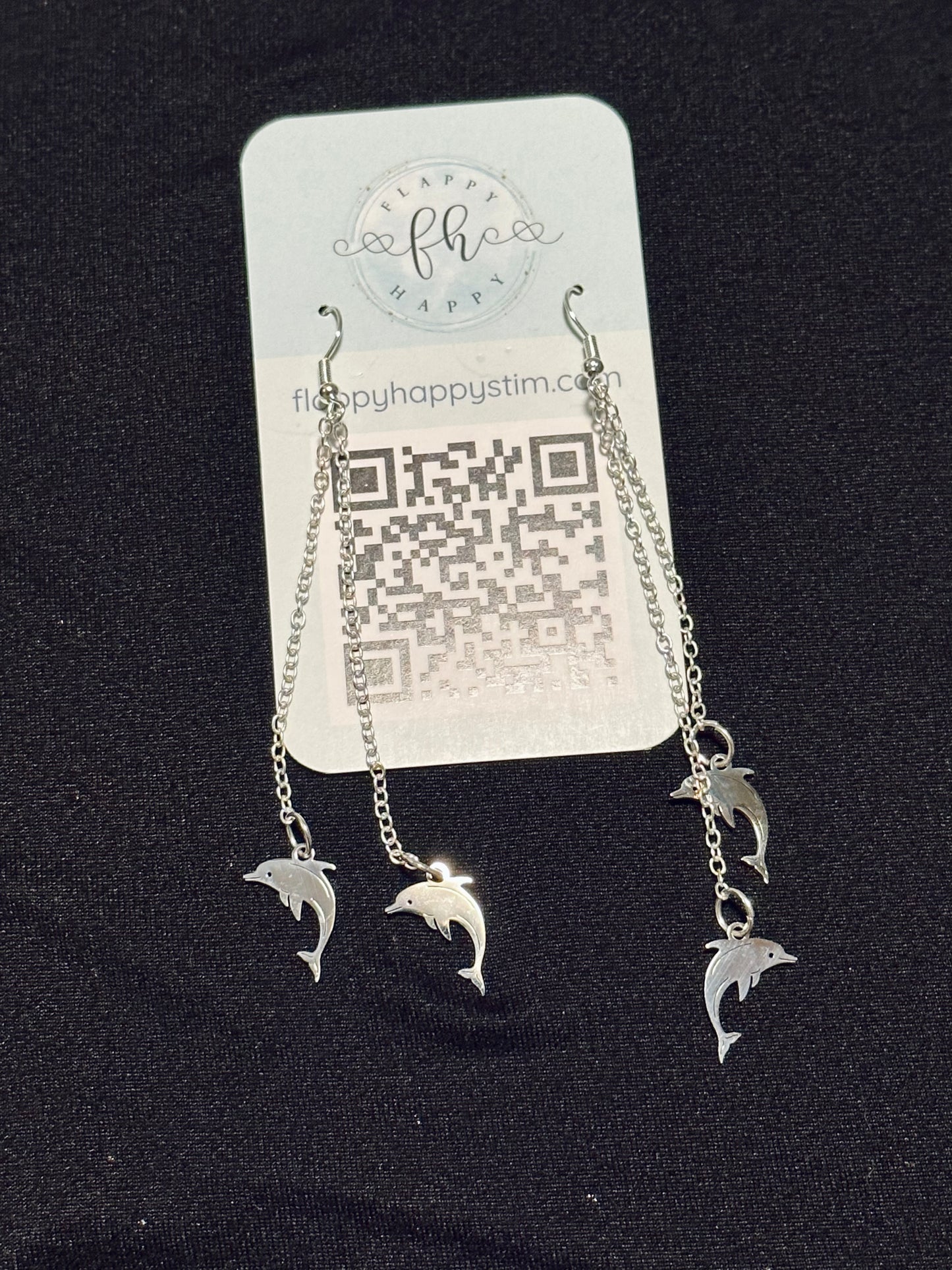 A pair of silver fidget earrings on a blue business card. The earrings have fishhook ends with two dangling chains on each hook. At the end of the chains are silver dolphin charms. The earrings are designed to be pulled back and forth.