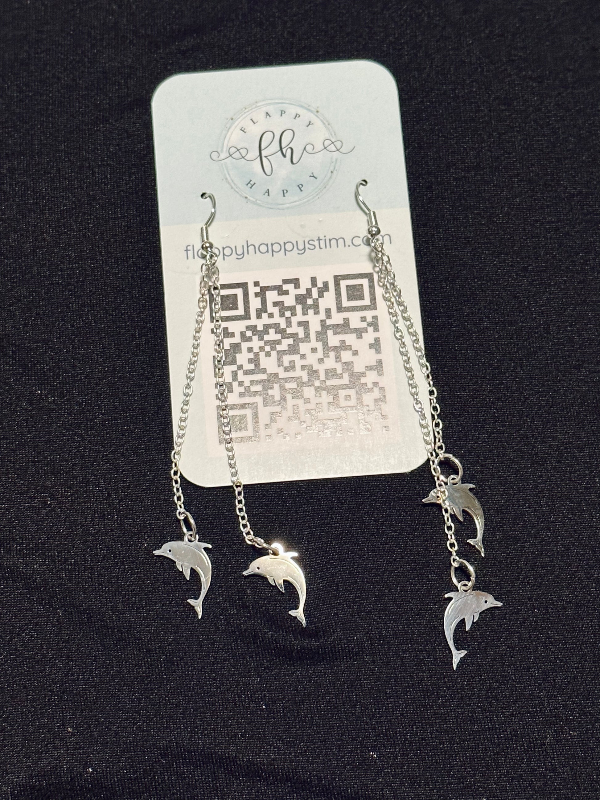 A pair of silver fidget earrings on a blue business card. The earrings have fishhook ends with two dangling chains on each hook. At the end of the chains are silver dolphin charms. The earrings are designed to be pulled back and forth.