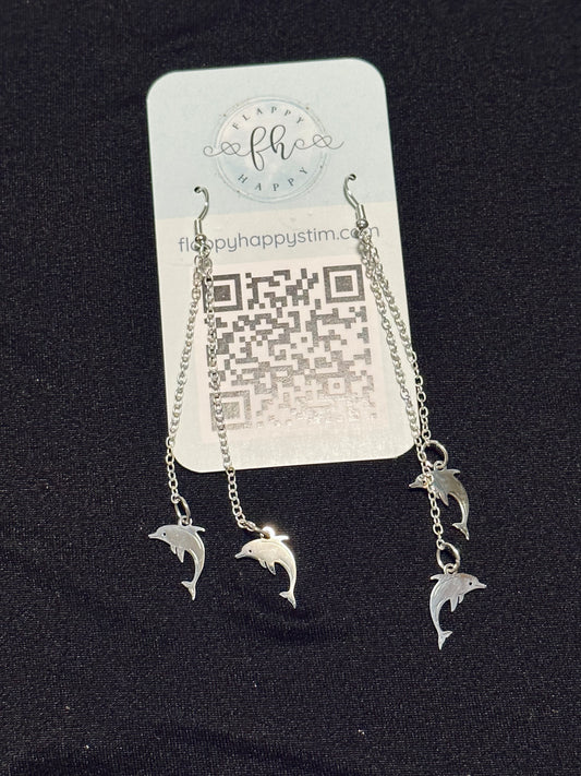 A pair of silver fidget earrings on a blue business card. The earrings have fishhook ends with two dangling chains on each hook. At the end of the chains are silver dolphin charms. The earrings are designed to be pulled back and forth.