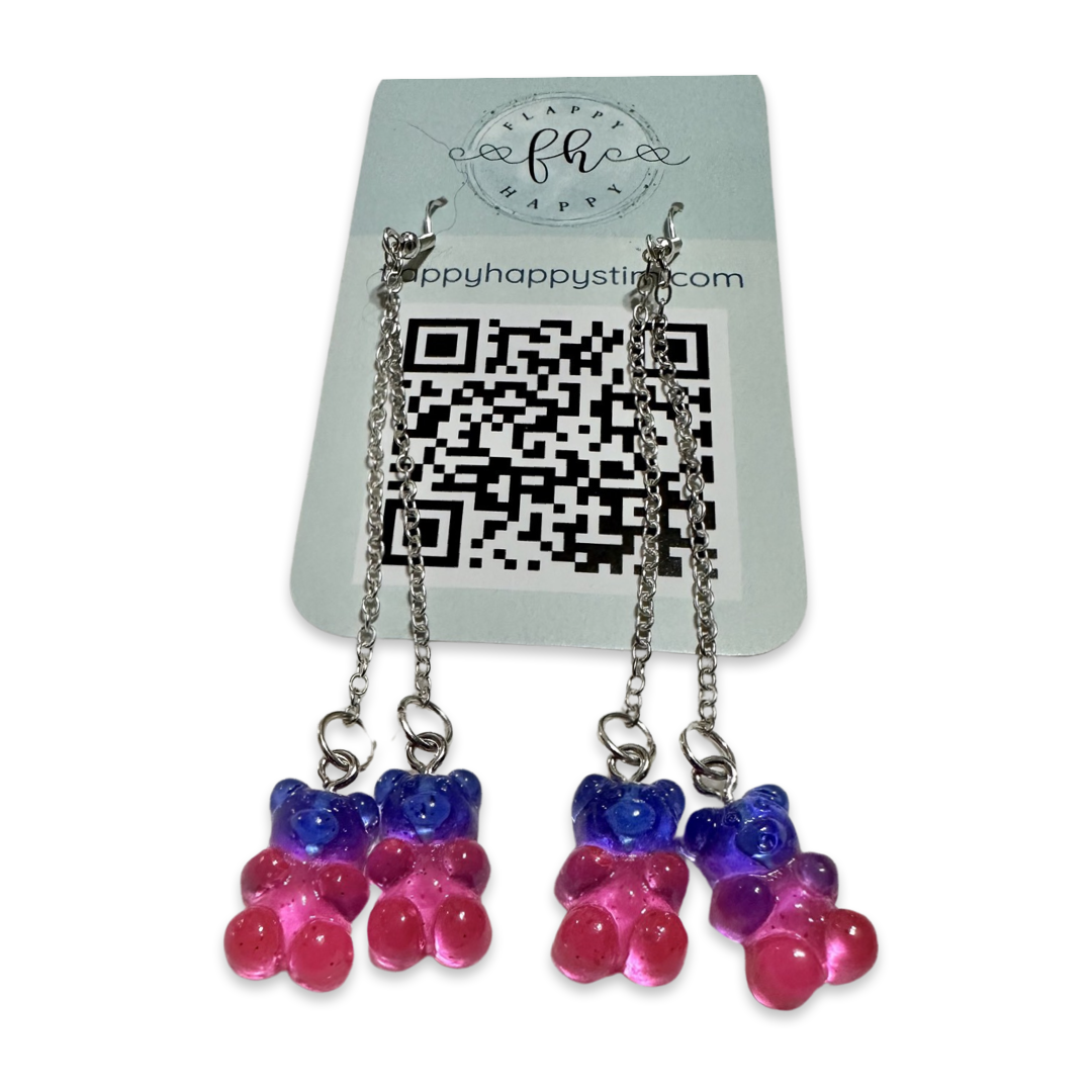 A pair of fidget earrings on a blue business card. The earrings have fishhook ends with two dangling chains on each hook. At the end of the chains are charms that look like dark blue and pink gummy bears. The earrings are designed to be pulled back and forth.