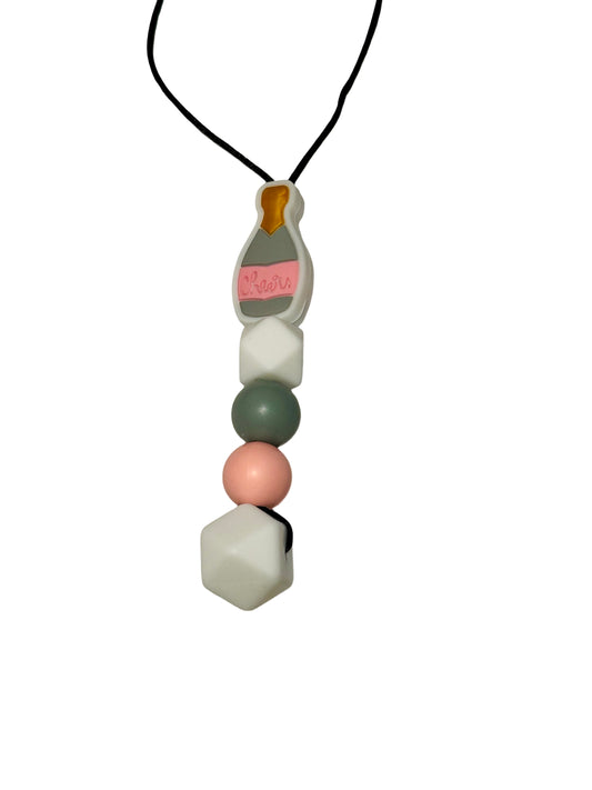 There is a bead chewable necklace. The top bead is a champagne bottle that says ‘cheers’ on it. It is grey and pink. Underneath are four more beads. A white hexagon, a grey round bead, a pink round and a large white hexagon.