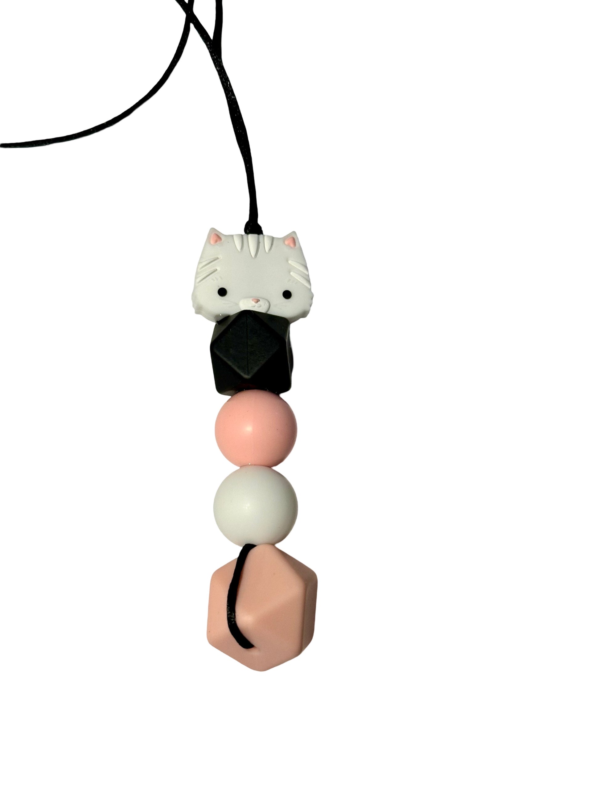 There is a cat chewable bead at the top of this chewable bead necklace. It is followed by a black hexagon, a pink round bead, a white round bead and a larger pink hexagon.