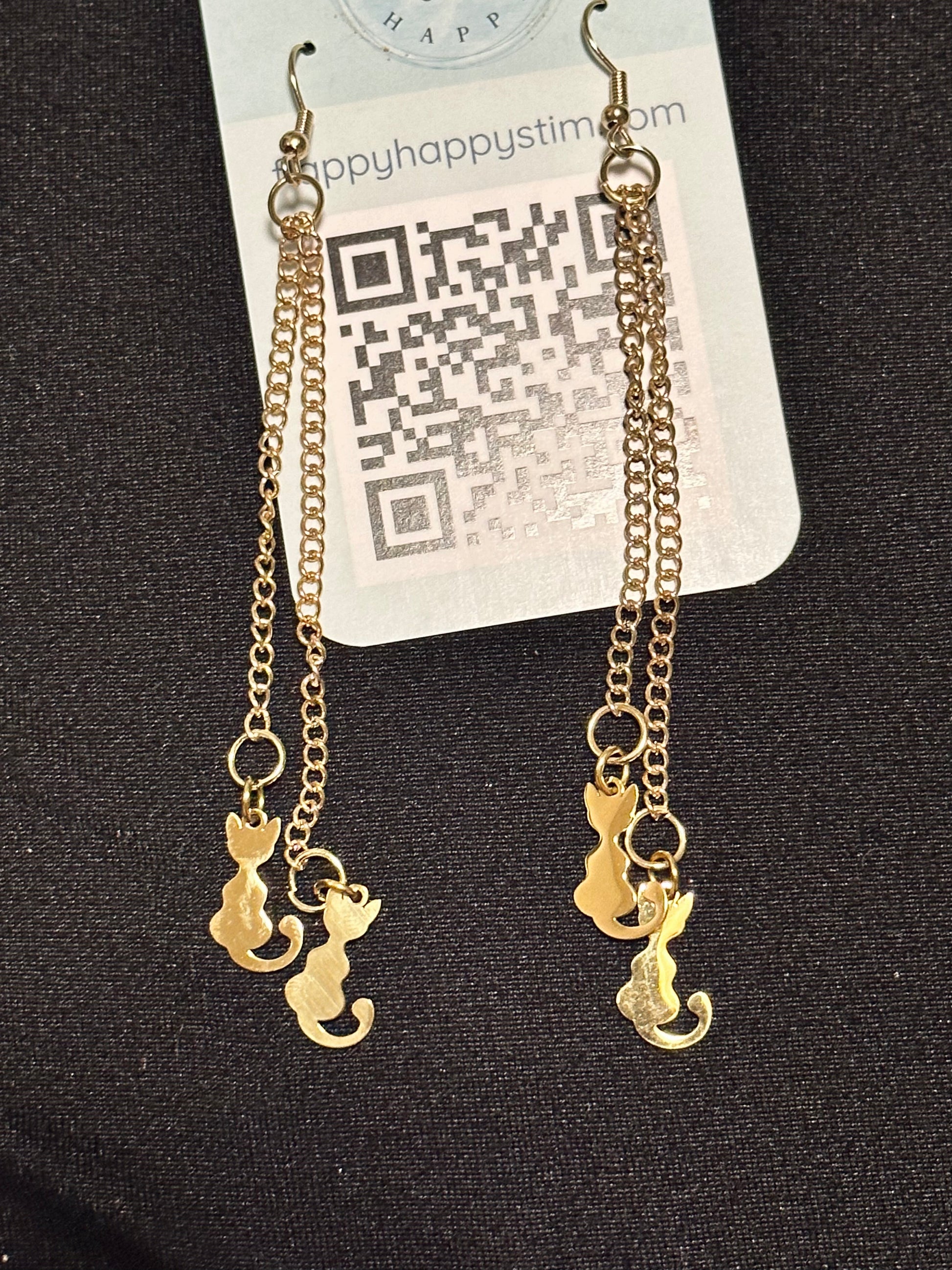 A pair of gold fidget earrings on a blue business card. The earrings have fishhook ends with two dangling chains on each hook. At the end of the chains are gold cat charms. The earrings are designed to be pulled back and forth. 