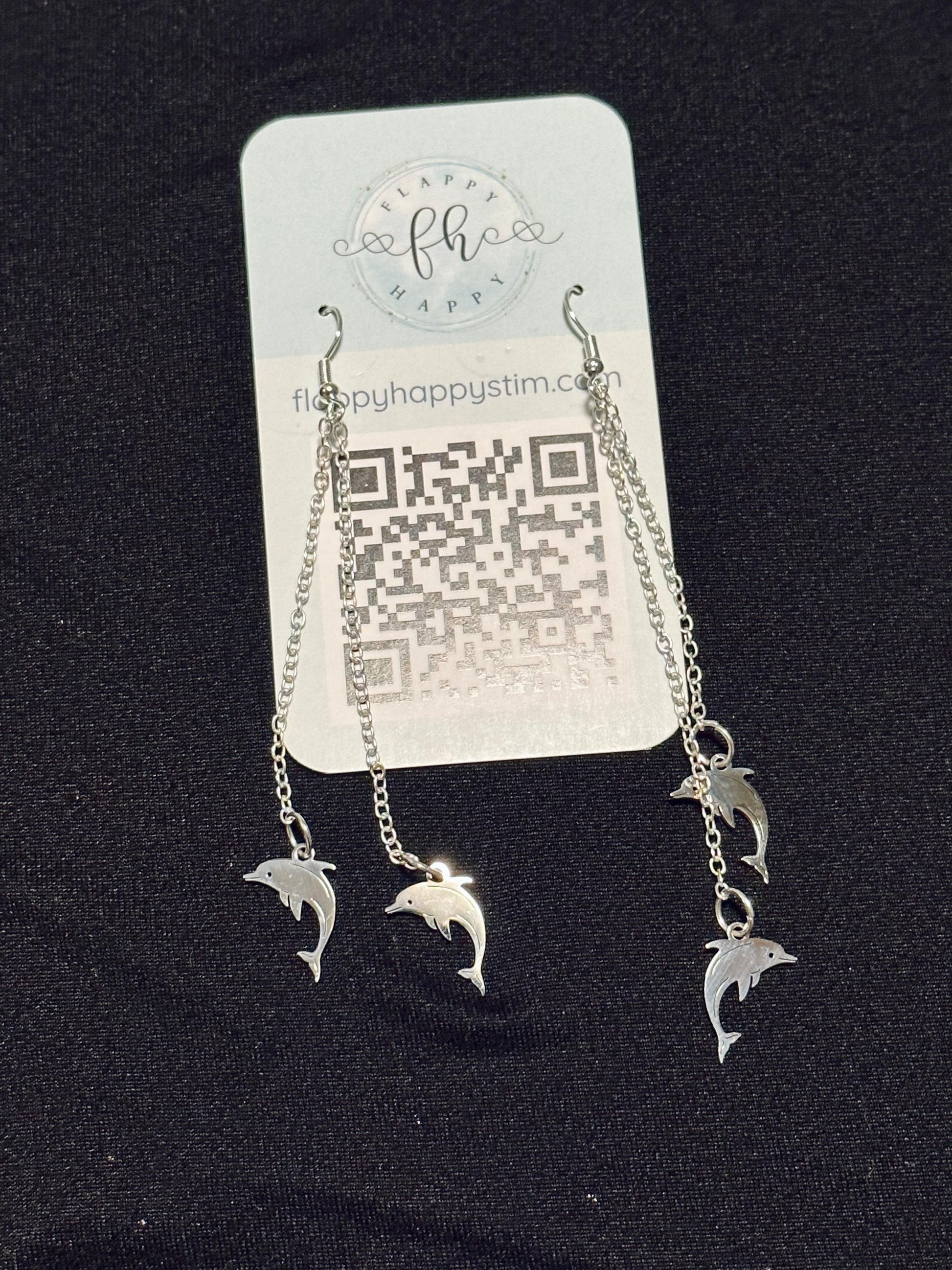 A pair of silver fidget earrings on a blue business card. The earrings have fishhook ends with two dangling chains on each hook. At the end of the chains are silver dolphin charms. The earrings are designed to be pulled back and forth. 