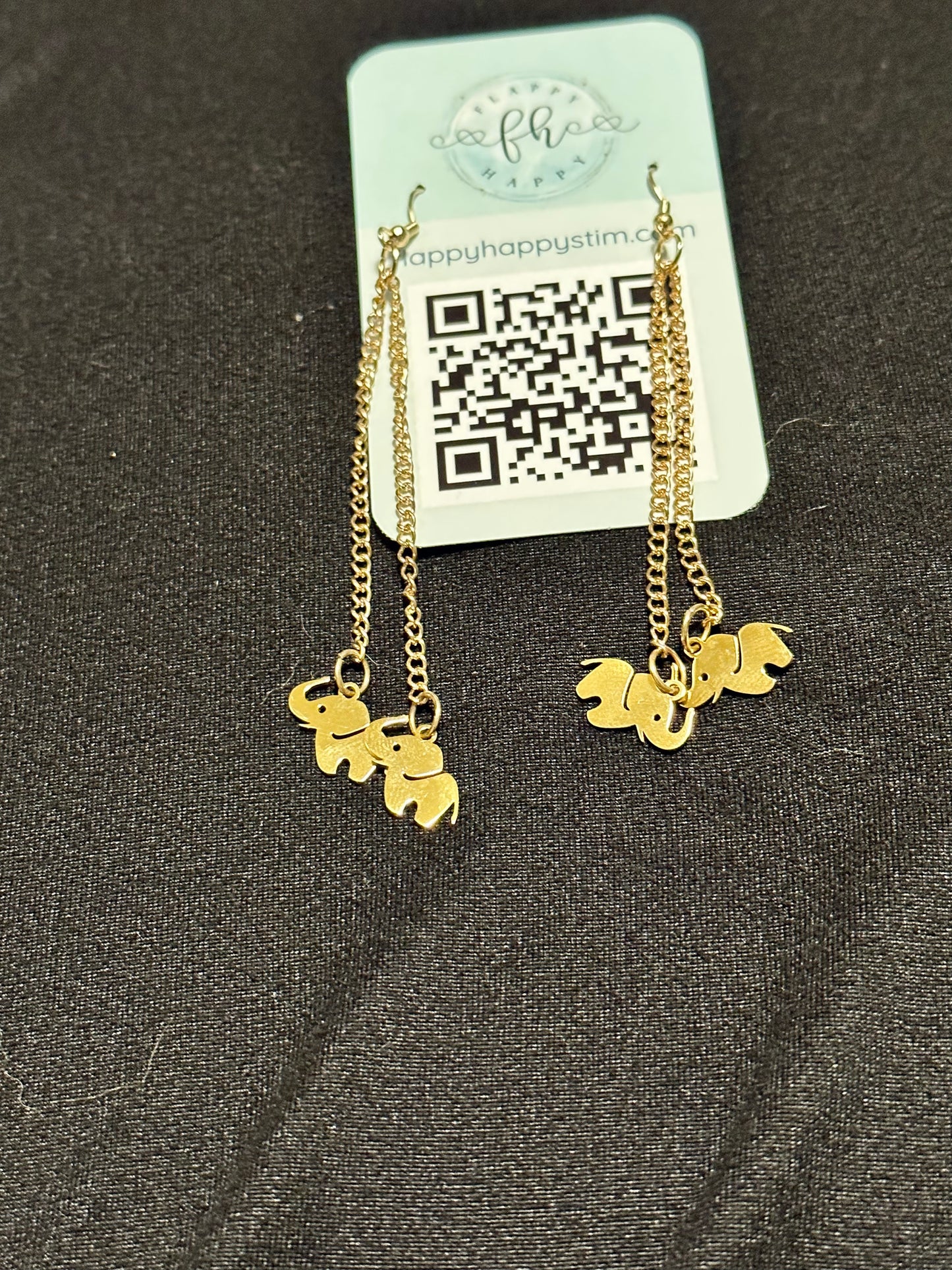 A pair of gold fidget earrings on a blue business card. The earrings have fishhook ends with two dangling chains on each hook. At the end of the chains are gold elephant charms. The earrings are designed to be pulled back and forth. 