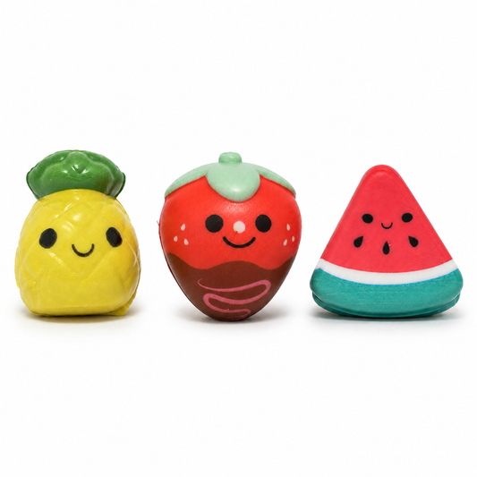 Three smiling fruit squishies displayed against a white background: a yellow pineapple, a chocolate-covered strawberry with pink drizzle, and a triangular watermelon slice.