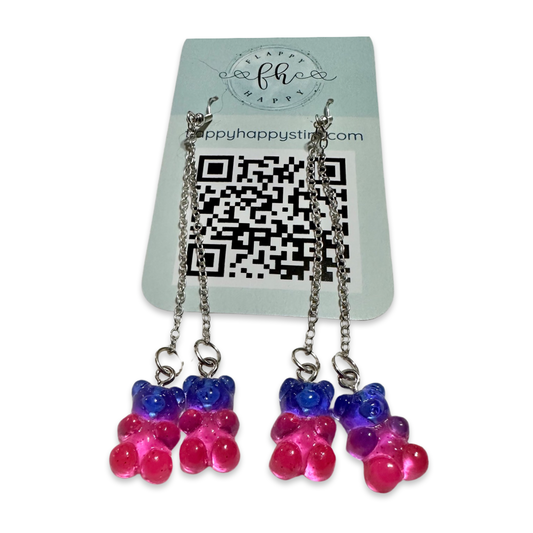 A pair of fidget earrings on a blue business card. The earrings have fishhook ends with two dangling chains on each hook. At the end of the chains are charms that look like dark blue and pink gummy bears. The earrings are designed to be pulled back and forth. 