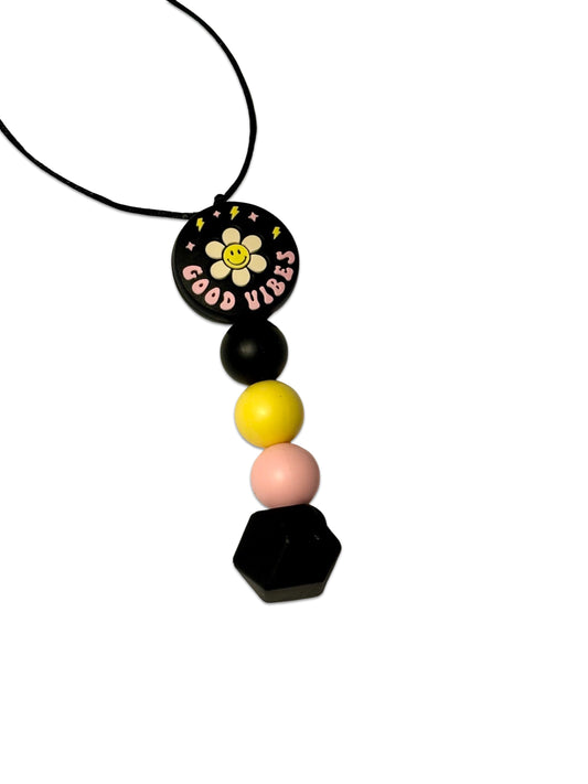 There is a black circle bead at the top of the bead chewable necklace. It has a flower on it with the words ‘good vibes’ underneath. There are four beads underneath this one. They are a black round bead, a yellow round bead, a pink round bead and a black hexagon.