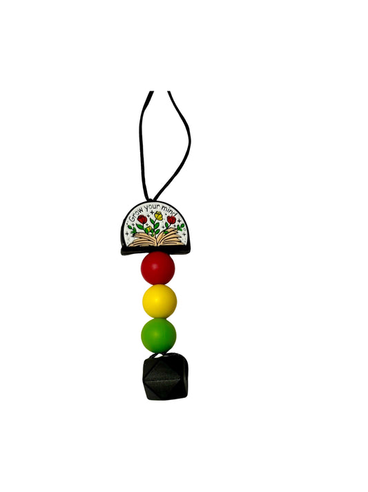 A chewable bead necklace with a half circle shaped bead at the top. It says “grow your mind” and shows flowers coming out of a book. There is then a red round bead, a yellow round bead, and a green round bead followed by a large black hexagon bead. 