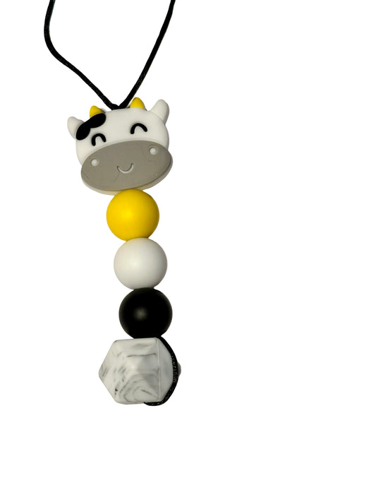 There is a cow bead at the top of this chewable bead necklace. There are three round beads underneath in the following colours: yellow, white and black. There is a grey and white speckled hexagon at the bottom. 