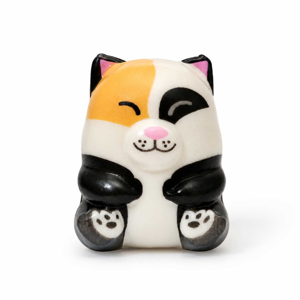 A cute, stylized cat figurine with a tri-color calico pattern sitting in a crouched pose with its paws together. It has a pink nose and inner ears, and black paw-print details on its feet, set against a plain white background.