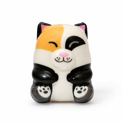 A cute, stylized cat figurine with a tri-color calico pattern sitting in a crouched pose with its paws together. It has a pink nose and inner ears, and black paw-print details on its feet, set against a plain white background.