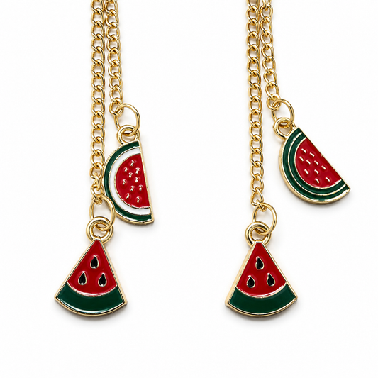 Close-up of a pair of gold-tone chain earrings, each featuring one triangular watermelon charm and one rounded watermelon-slice charm in red, green, white, and gold.