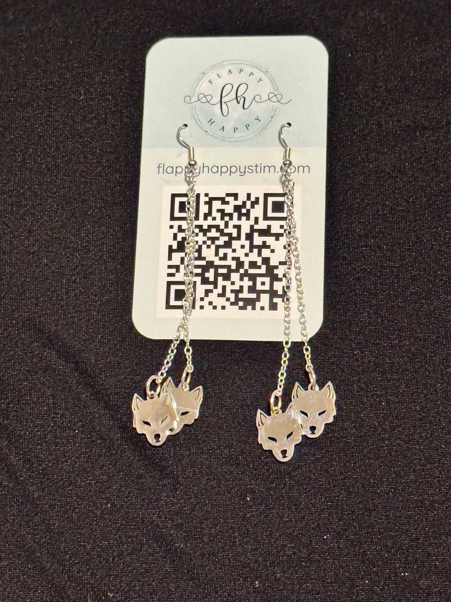 A pair of silver fidget earrings on a blue business card. The earrings have fishhook ends with two dangling chains on each hook. At the end of the chains are silver wolf head charms. The earrings are designed to be pulled back and forth. 